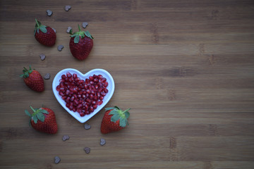 healthy food photography image of fresh red strawberries with green leaf top and shiny fruit pomegranate seeds placed in a white love heart shape dish with natural dark wood background and copy space