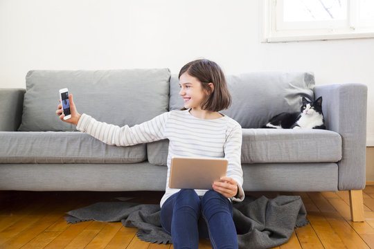 Happy girl with tablet sitting on the floor of the living room taking selfie with smartphone