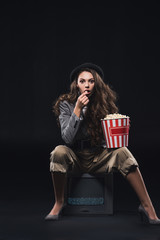 Naklejka premium shocked young woman eating popcorn while sitting on tv and looking at camera on black