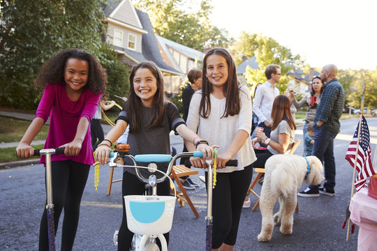 Three Pre-teen Girls On Scooters And A Bike At A Block Party