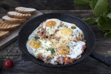 Fried eggs in an old frying pan with cheese, tomatoes, lettuce leaves and fried bread on a wooden background.