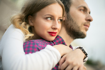 Portrait of couple of young female with blonde wavy hair with red lips and in white top  and young male in checked shirt with beard embracing outdoors /close-up portrait of young couple embracing