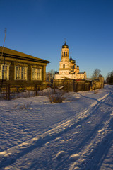 Russian village in the winter. Peasant old traditional wooden house and the Church of the Nativity of Christ on a winter snowy street at sunset.