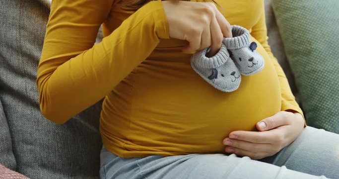 Close up of the big pregnant belly and woman's hands playing with small kid's sleepers near it. on the gray sofa. Inside