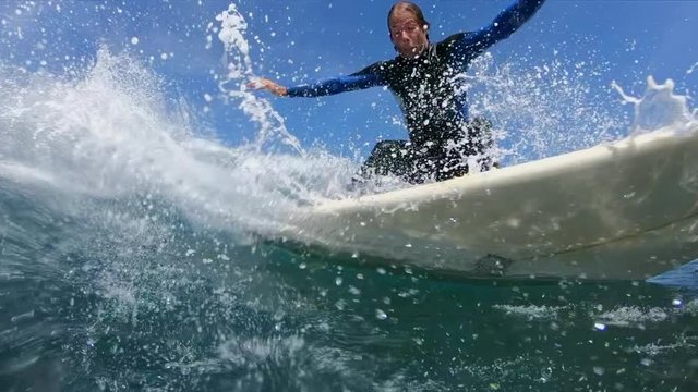SLOW MOTION, LOW ANGLE: Surfer guy makes a sharp turn on his surfboard and splashes water on camera. Cheerful surfboarder looks back after carving a big tube wave in on beautiful sunny day in nature.