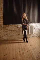 The girl hesitates and stands at the battery, a brick wall and a large window hung with curtains. Full length portrait of blonde girl wearing black leather outfit.