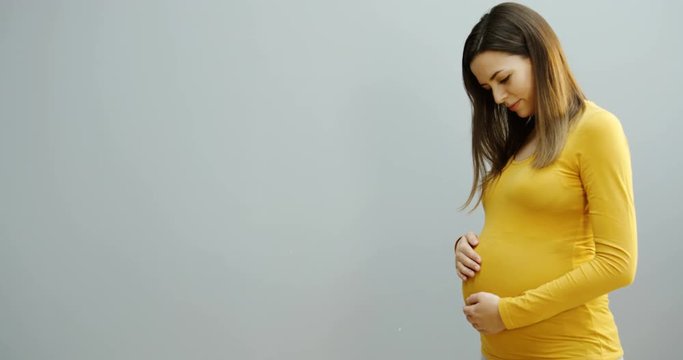 Portrait shot of the caucasian good looking pregnant woman in a yellow sweater standing a half turn to the camera on the white wall background and stroking her big belly. Indoors