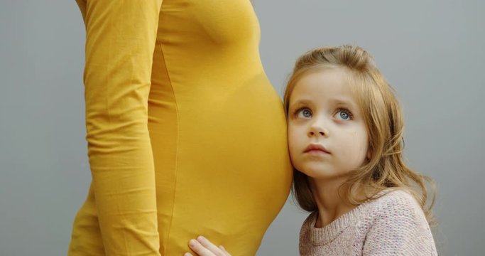 Portrait shot of the pretty small girl putting her ear at the pregnant belly and than looking up and kissing it on the gray wall background. Close up. Indoors