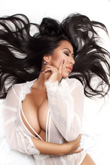 Naklejka premium Beautiful brunette is lying on the bed, in a room. She is wearing a white bodysuit and high hells. Mixed race Asian / Caucasian female model. Glamour and boudoir photography.