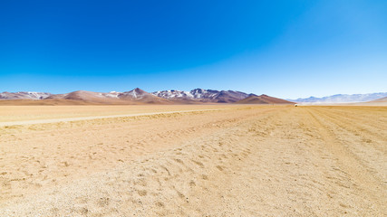 Fototapeta na wymiar Dirt road at high altitude with sandy desert and barren volcano range on the Andean highlands. Road trip to the famous Uyuni Salt Flat, travel destination in Bolivia.