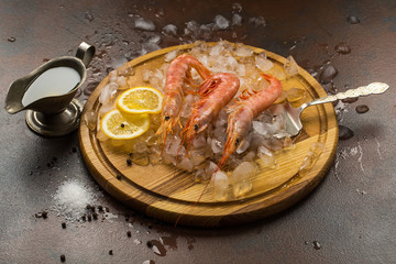 Large tiger Shrimps or langostinos with lemon on ice on wooden cutting board. Cutler, sauce on dark background