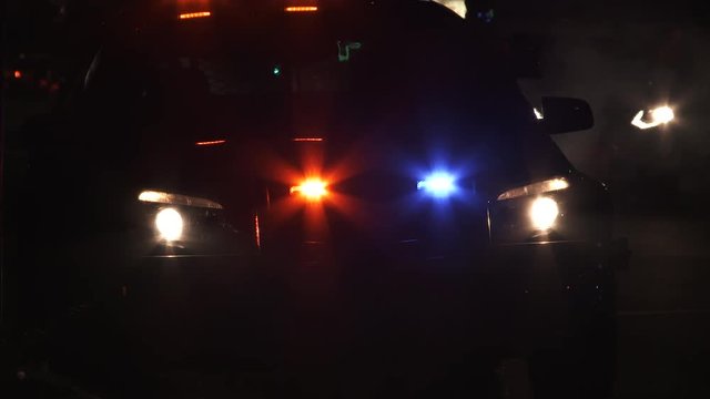 Unrecognizable people walking passed police car with flashing lights at night.