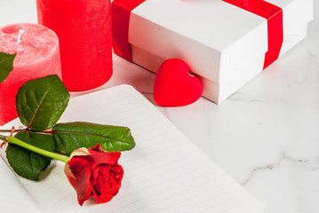 Holiday  background, Valentine's day. Bouquet of red roses, tie with a red ribbon, with blank notepad, wrapped gift box and red candle. On a white marble table, copy space top view