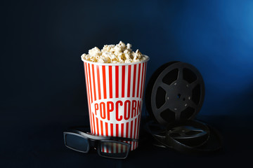 Paper cup with tasty popcorn, glasses and movie reel on dark background