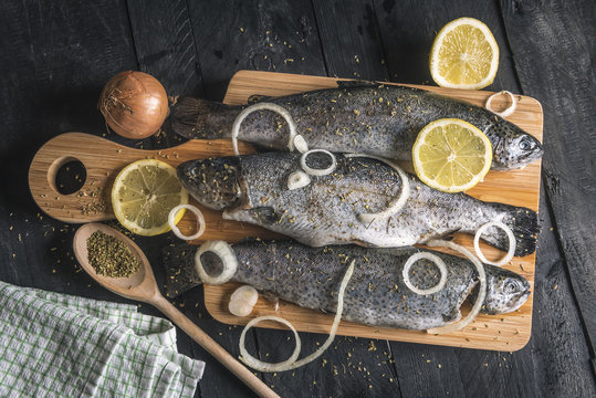 Fresh fish seasoned with herbs and lemons - High angle view with uncooked fish on a wooden cutting board, seasoned with dried herbs, lemon slices and onion rings, on a vintage black table.
