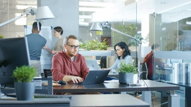 Busy International Corporate Office, Caucasian Man Working at His Desk on a Laptop. In Background Creative Young People Working.  Shot on RED EPIC-W 8K Helium Cinema Camera.