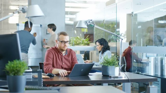 Busy International Corporate Office, Caucasian Man Working at His Desk on a Laptop. In Background Creative Young People Working.  Shot on RED EPIC-W 8K Helium Cinema Camera.