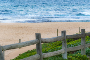 Scenic view of abandoned sandy beach and ocean waves, wooden fence in foreground. Empty Mona Vale...