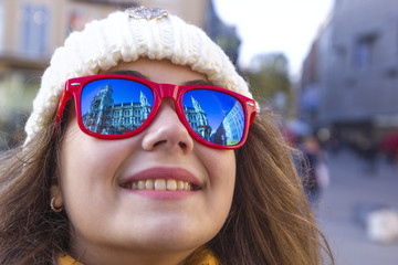 Portrait of a cheerful young girl close up. The New Town Hall of Munich is reflected in the red sunglasses.
