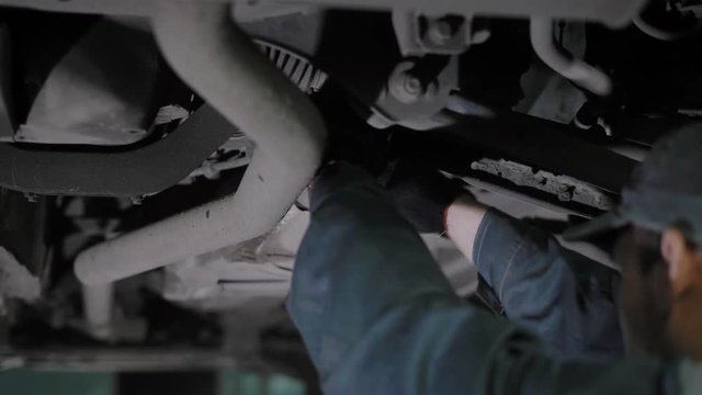 automobile mechanic is regulating handbrake cable, standing under lifted car, tightens details in a car repair workshop