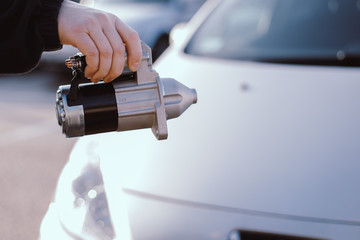 A man hand holding an electric starter with a car in the background, car spare parts.