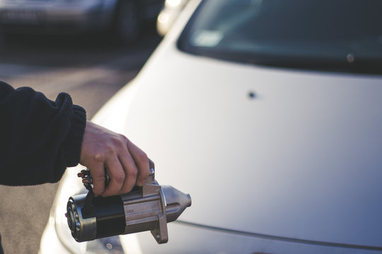 A man hand holding an electric starter with a car in the background, car spare parts.