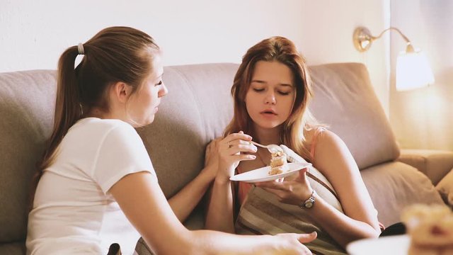 Portrait of cheerful females talking and eating cake at home on sofa
