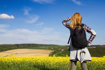 Young hiking woman is looking out over the countryside. Beautiful yellow rape field and blue sky in the background.