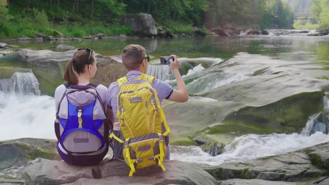 A young couple of tourists admiring the mountain river, a man taking pictures of the river with a smartphone