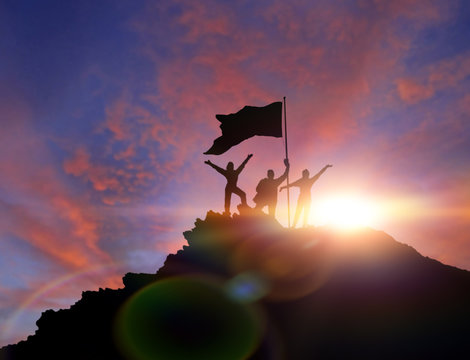 High Achiever, Silhouettes Of Three People Holding On Top Of A Mountain To Raise Their Hands Up. A Man On Top Of A Mountain. Conceptual Design. Against A Dramatic Sky With Clouds At Sunset.