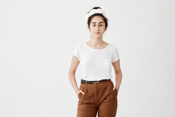 Cute pretty teenage girl wears loose white t-shirt and brown trousers, holds her hands in pocket, feels enjoyment. Pleasant-looking girl posing against white background
