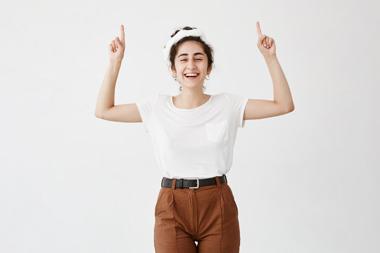 Waist up shot of joyful happy girl wearing white t-shirt looking at camera, pointing fingers at copy space above her head. Young woman indicating something on blank studio wall with hands