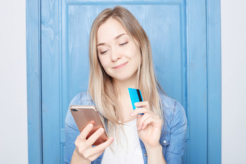 Horizontal portrait of blond female has dreamful expression, imagines new clothes, going do online shopping, holds modern cell phone and plastic card, isolated over white background with copy space.