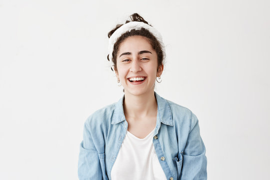 Dark-haired positive girl wears do-rag and denim shirt, looks happily at camera, smiles broadly, laughs at joke, rejoices youth. Female model poses against white backgorund demostrates white teeth