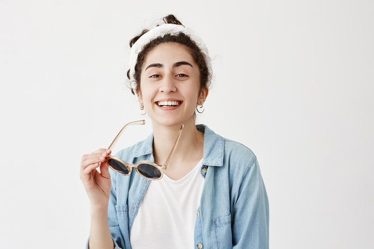 Joyful dark-haired female student in do-rag smiles broadly, demonstrates white even teeth, dressed in denim shirt, has great day, relaxing indoors isolated against white background with copy space.