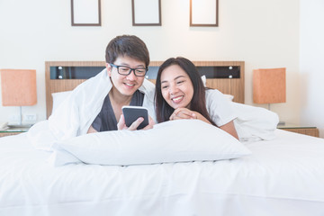 Asian couple using mobile phone on white bed in morning time