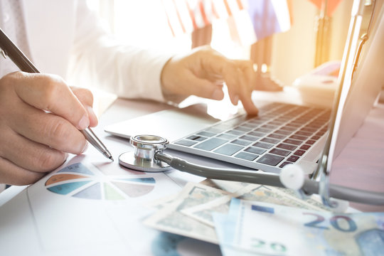 Doctor Working On Laptop Computer With Report Analysis And Money About Healthcare Costs And Fees In Medical Hostpital Office. Focus Stethoscope On Table. Healthcare Budget And Business Concept