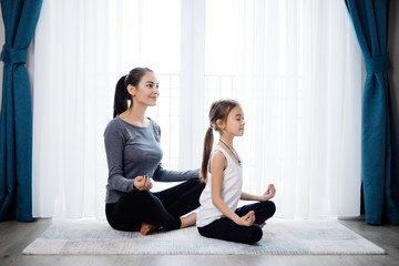 Beautiful young woman and her charming little daughter are smiling while doing yoga together at home
