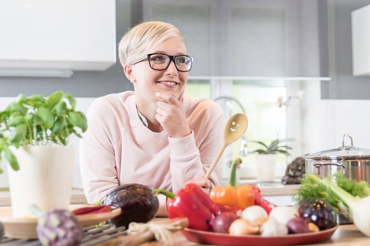 Junge h&uuml;bsche Frau in ihrer K&uuml;che l&auml;chelt und &uuml;berlegt, welches Gem&uuml;se sie kochen soll