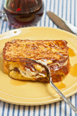 Freshly baked, maple bacon stuffed french toast with hot maple syrup and artisan knife and fork on a round, yellow plate with glass vessel of syrup on a blue and white stripped towel