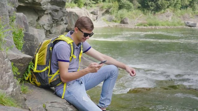 A young man with a yellow backpack is sitting near a mountain river. Uses a smartphone. Tourism and Technology Concept