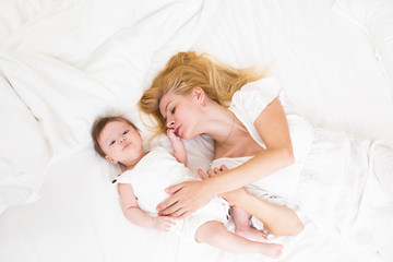 Mother and child on a white bed top view. Mom and baby girl with blue eyes playing in white bedroom. Parent and little kid relaxing at home and having fun together