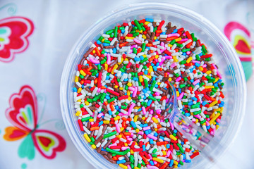 a bowl full of colorful candy sprinkles with a clear plastic spoon in them and a plastic tablecloth