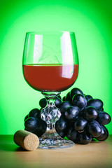 A glass of red wine next to bunch of black grapes and a cork on a wooden table on a green background (concept)