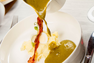 Cream of sorrel and green vegetables soup with quail egg, sour cream, mashed potatoes and crispy bacon being poured from a jug