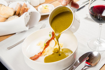 Cream of sorrel and green vegetables soup with quail egg, sour cream, mashed potatoes and crispy bacon being poured from a jug