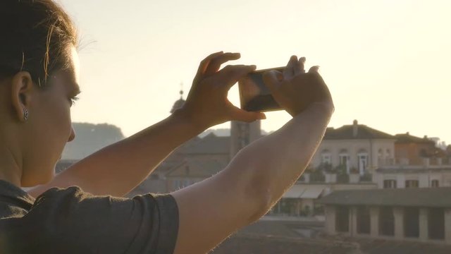 Beautiful young woman on high balcony in campidoglio taking pictures with smartphone of cityscape of Rome at sunset viewing historic buildings and domes slow motion steadycam