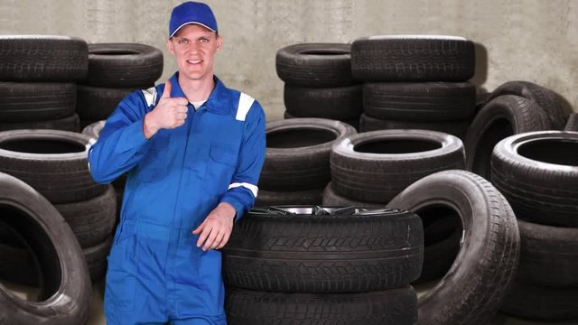 Caucasian car mechanic showing thumb up with piles of tires on the background