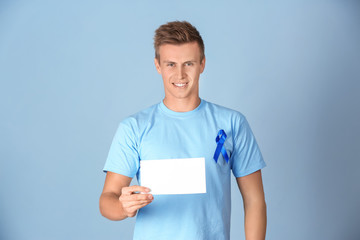 Young man in t-shirt with blue ribbon holding blank card on color background. Prostate cancer awareness concept