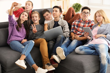 Obraz premium Group of teenagers with modern devices taking selfie while sitting on sofa indoors